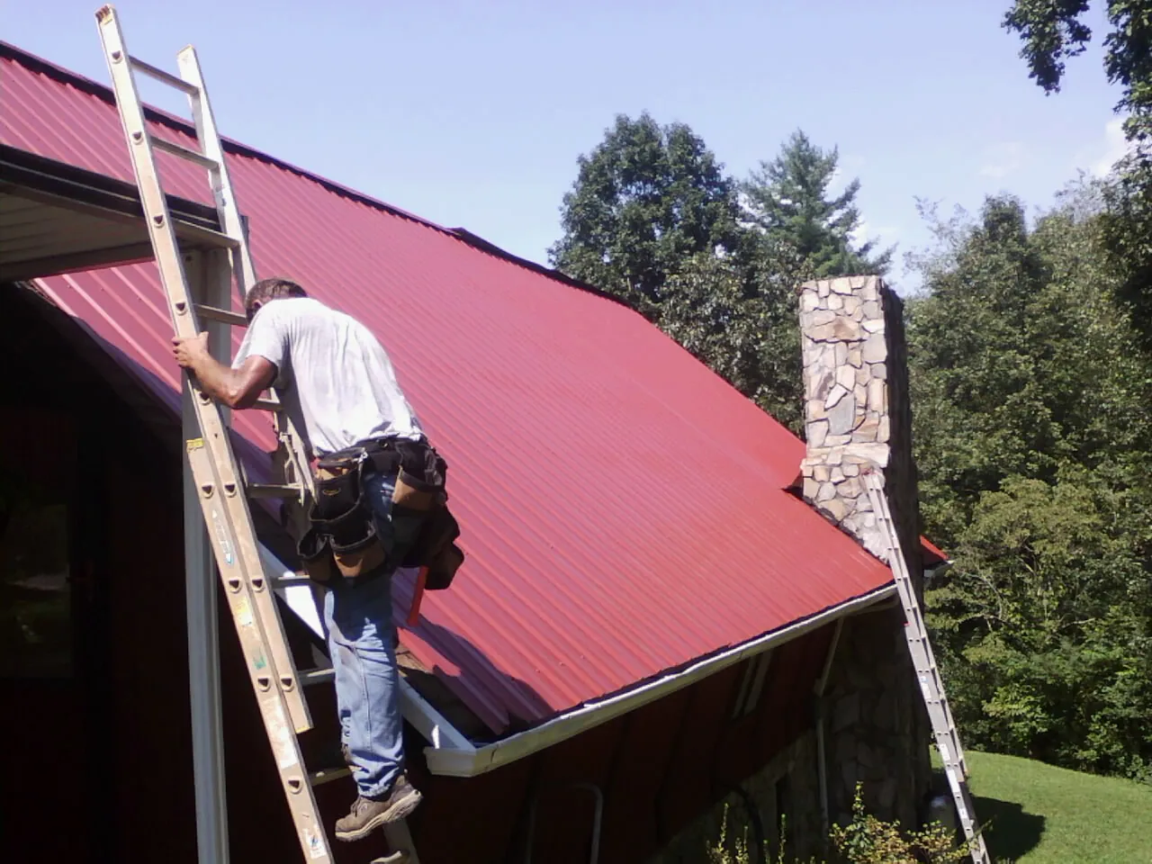 Roofer on a ladder beside a red metal roof and stone chimney — Soffit & Fascia Repair by Roofing Brothers in Otterbein